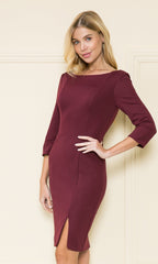 A woman models the Classy Knee-Length Career Dress 8526, a fitted burgundy dress with three-quarter sleeves and a front slit, while standing by a cream-colored wall.