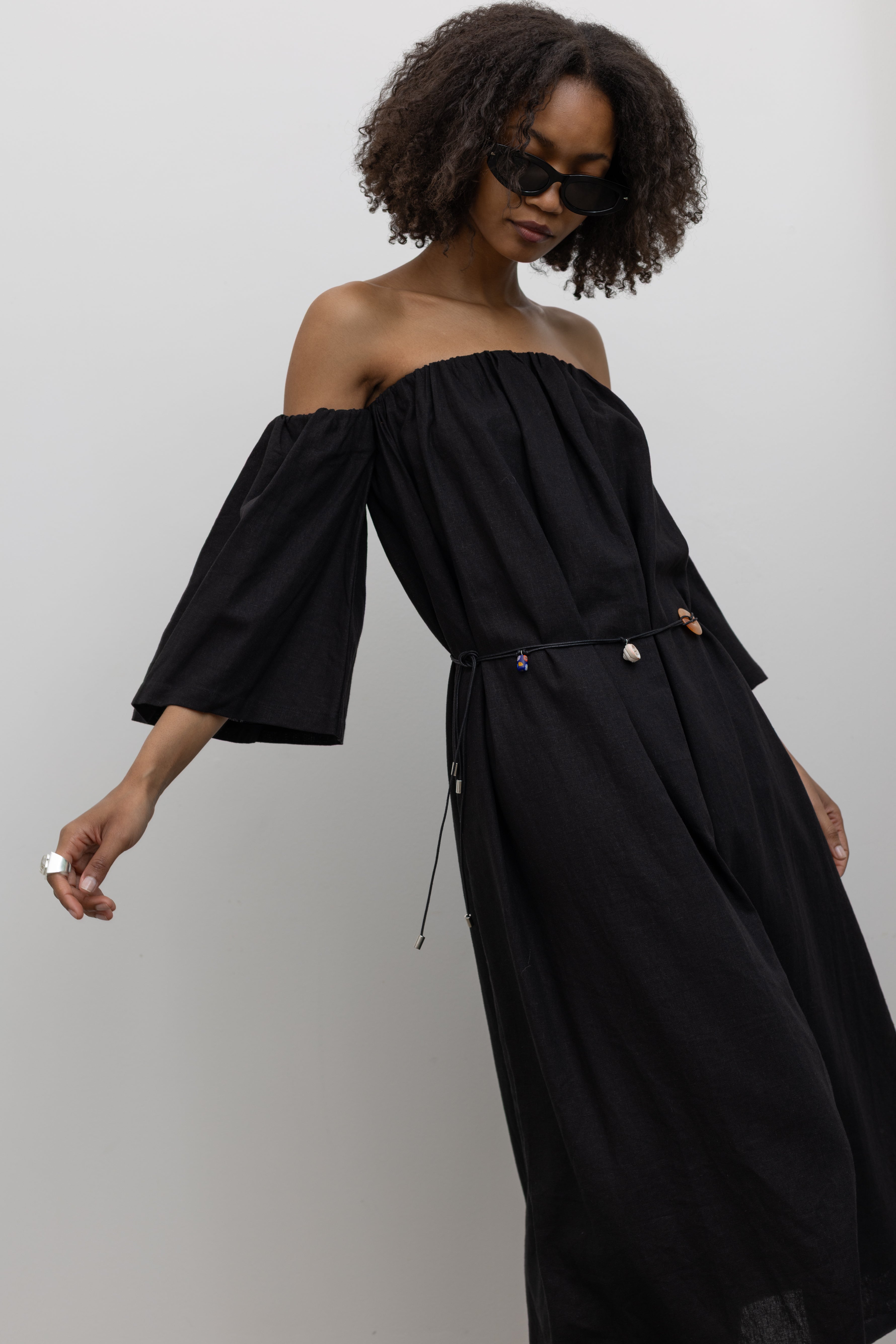 3/4 Front view of a woman in motion wearing a chic and versatile Alas Eius label cotton blend maxi dress featuring an elastic gathered top with removed flowy sleeves at an Alas Eius store.