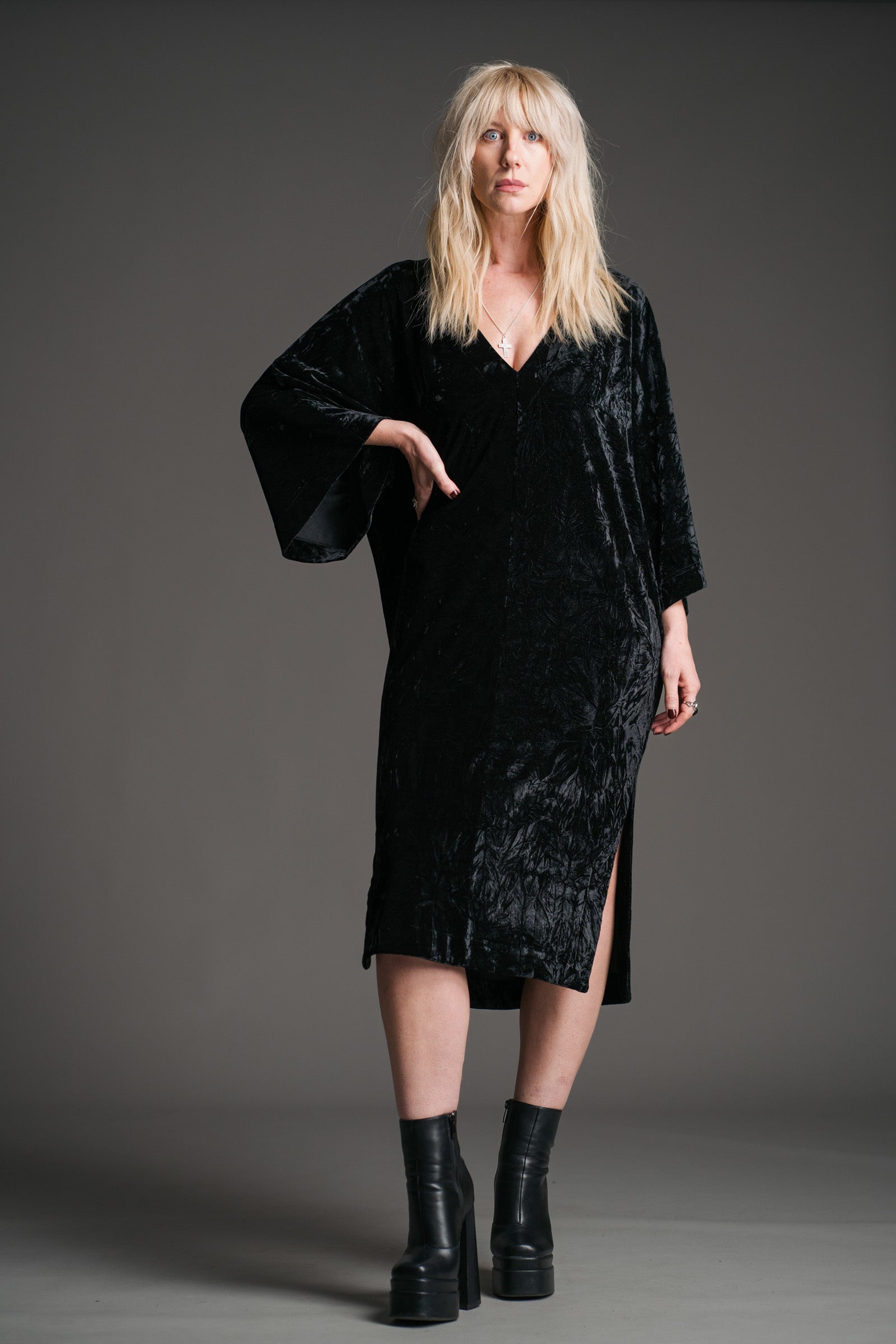 Sara wears Little Lies Rumours black velvet midi dress with wide sleeves and V‑neckline, styled with black platform boots in a studio full‑length front view.