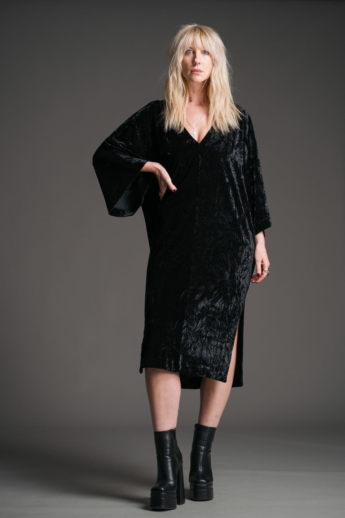 Sara wears Little Lies Rumours black velvet midi dress with wide sleeves and V‑neckline, styled with black platform boots in a studio full‑length front view.